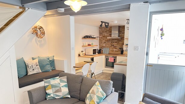 A modern living room with a grey sofa, a dining area, and a kitchen with a brick wall. - The Bolt Hole - a cottage that sleeps 4 guests in 2 bedrooms (St Ives)