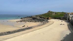 Une plage bordée d’une côte rocheuse, une colline herbeuse surmontée d’un bâtiment et un ciel dégagé.
