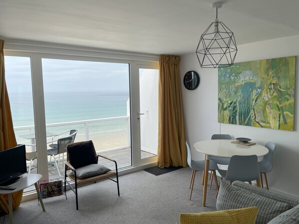 A modern room with a balcony overlooking the sea, a dining area with a round table, and a large abstract painting on the wall.