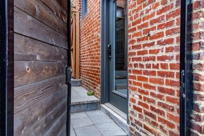 Un estrecho pasillo exterior con un muro de madera a un lado y un muro de ladrillo al otro, que conduce a una puerta con un panel de vidrio.