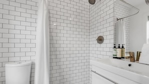 Une salle de bain avec des carreaux de céramique blancs rectangulaires, une douche, des toilettes, un lavabo avec un miroir et des articles de toilette posés sur le lavabo.