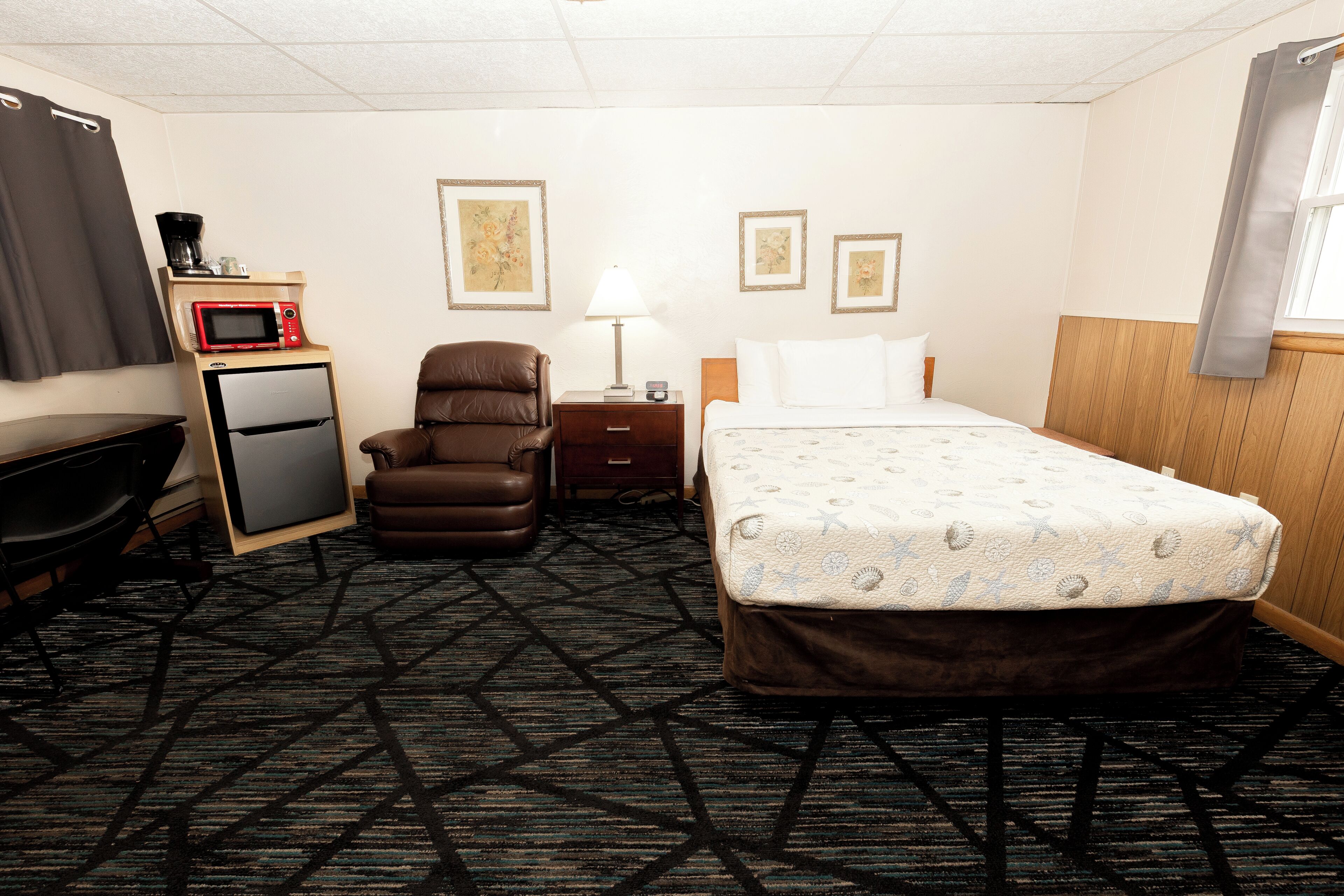 A hotel room with a bed, a desk, a chair, a microwave, a refrigerator, and a lamp.