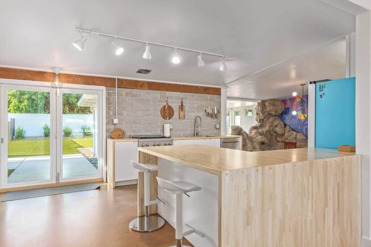 Une cuisine moderne avec un îlot central en bois, des tabourets de bar blancs et une grande fenêtre donnant sur un jardin.