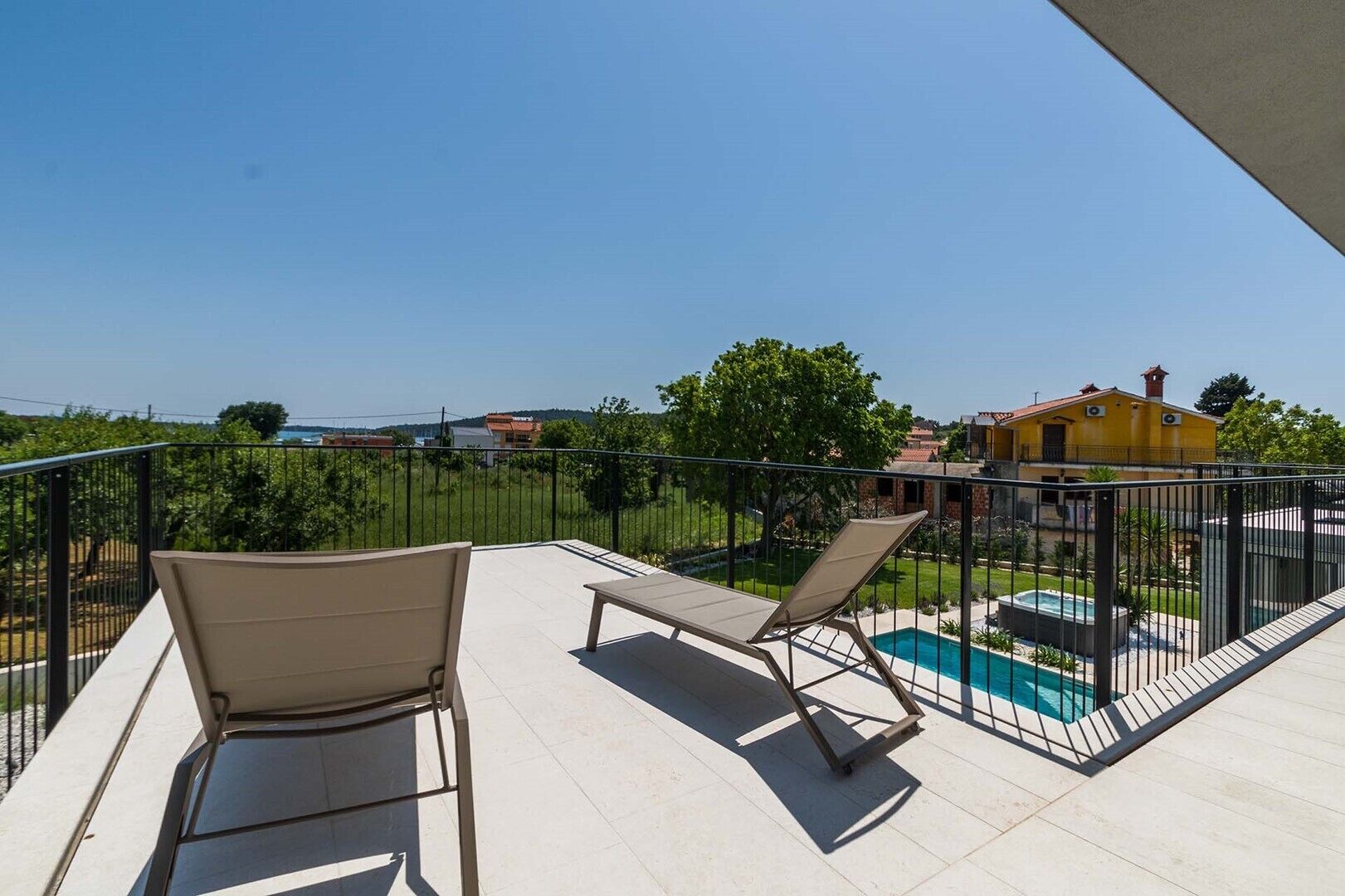 A balcony with two lounge chairs, a swimming pool, and a view of a yellow house and greenery.
