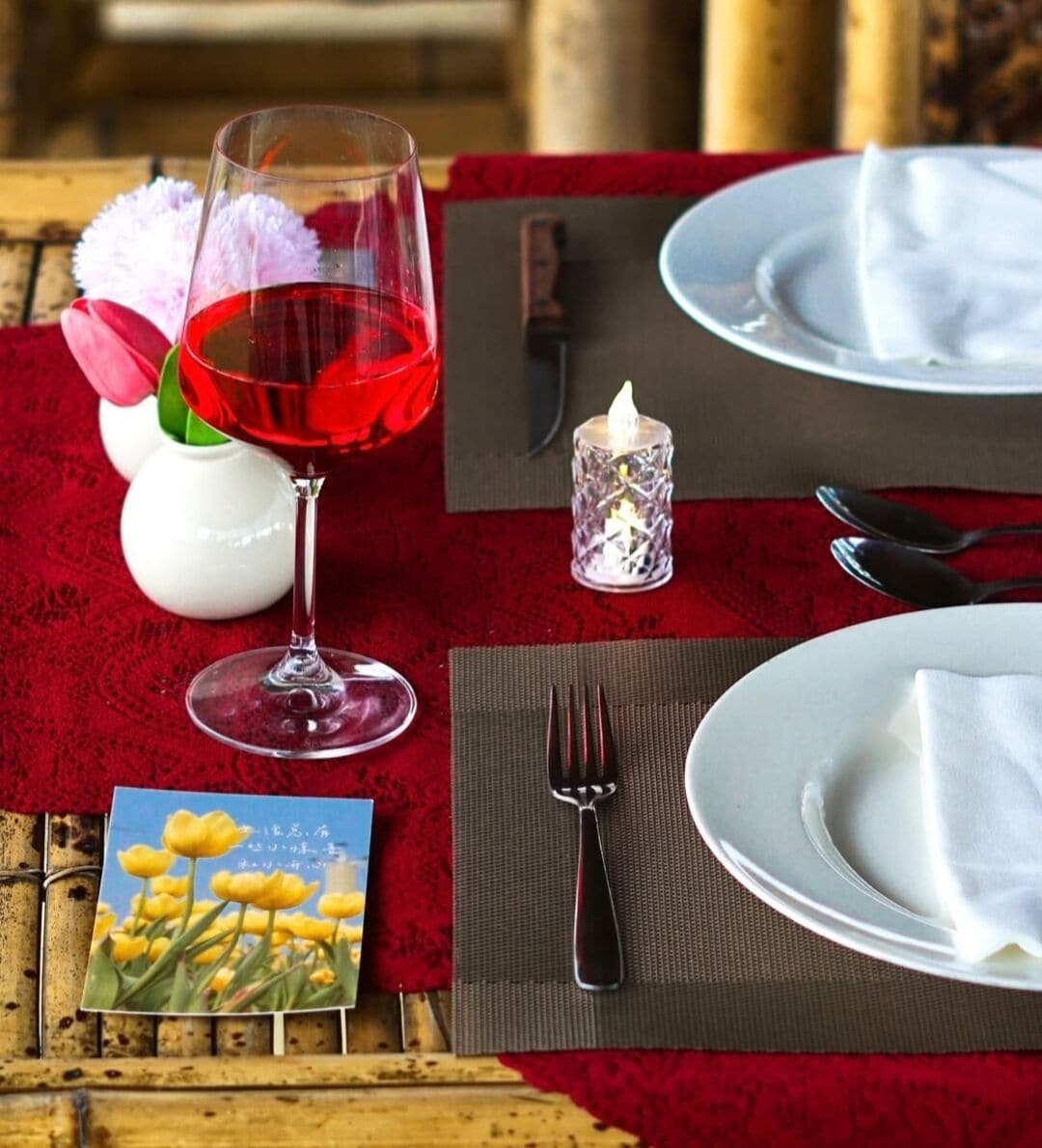 Une table dressée avec une nappe rouge, un verre de vin rouge, une bougie, une serviette et une petite carte ornée d’une image de fleur.