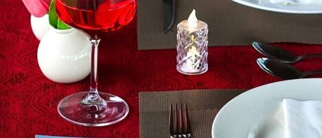 Une table dressée avec une nappe rouge, un verre de vin rouge, une bougie, une serviette et une petite carte ornée d’une image de fleur.