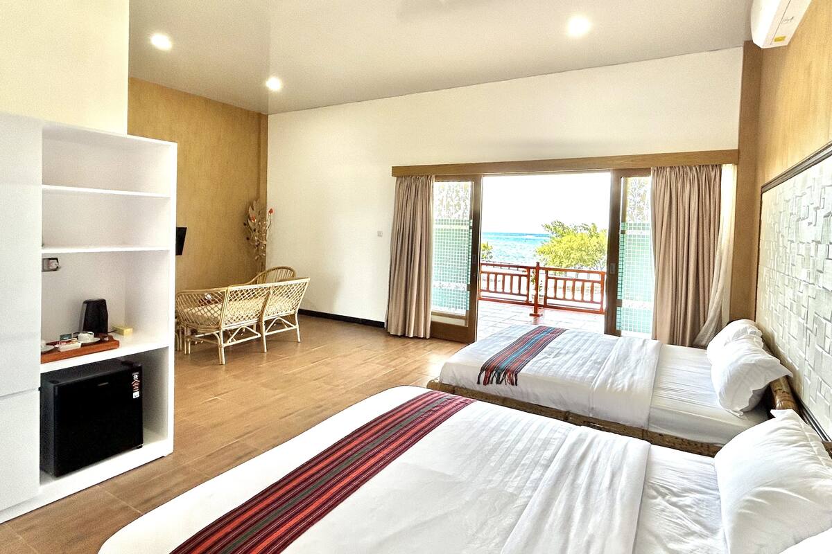 A hotel room with two beds, a small desk, a bookshelf, and a balcony with a view of the sea.