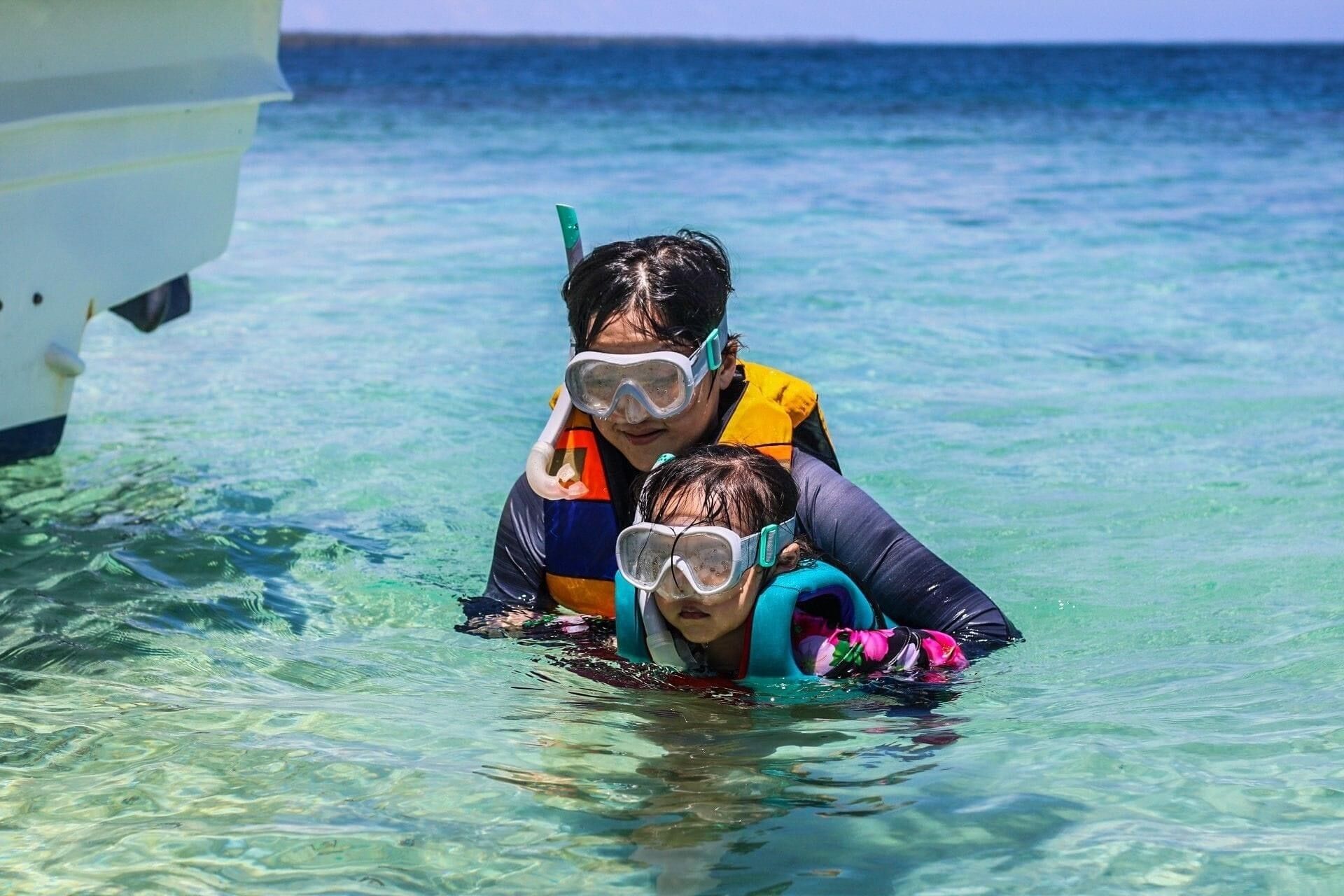 Dos personas haciendo snorkel en aguas azules cristalinas, con un bote al fondo.