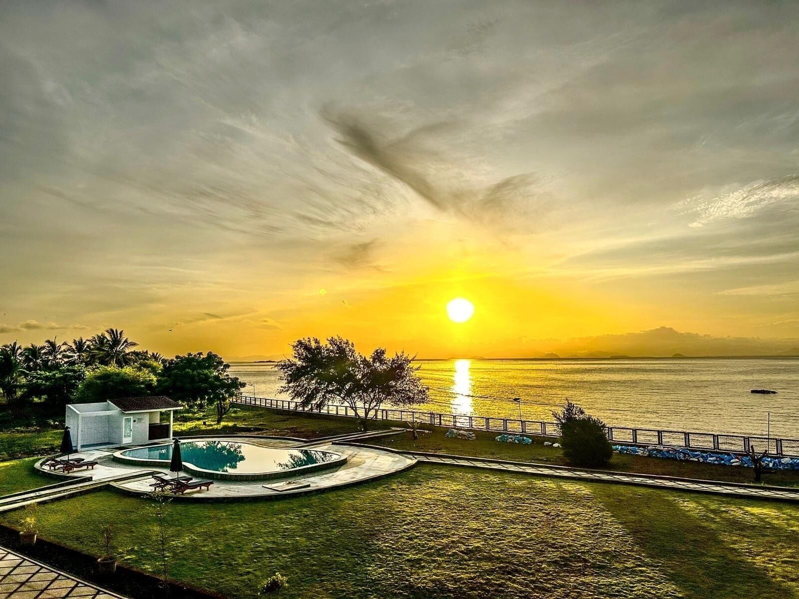 A pool area with a view of the sunset over the water, surrounded by a fence and palm trees.