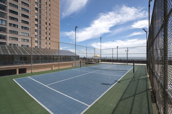 A tennis court with net, surrounded by a chain-link fence, and a tall building in the background.