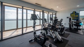 Un gimnasio con máquinas de cardio y equipo de pesas, ventanas amplias con vista al mar y un letrero que indica uso exclusivo para mayores de edad.