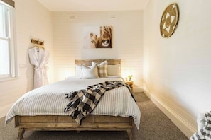 Un dormitorio con estructura de cama de madera, ropa de cama a rayas, cabecera con almohadas, una obra de arte en la pared de un león y una mesita de noche con lámpara.