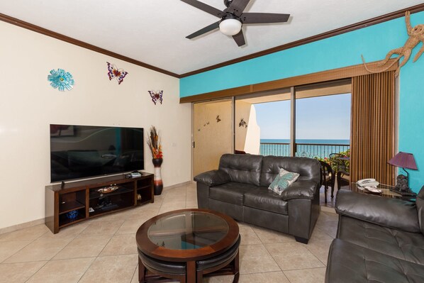 A living room with a TV, leather sofa, glass coffee table, and a balcony with a view.