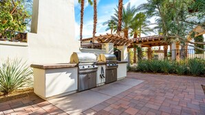 An outdoor kitchen with a grill and storage cabinets, surrounded by palm trees and a pergola. - LV029 Spacious Upstairs Studio near Pool/Hot Tub (La Quinta)