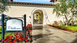 Entrada a un Clubhouse con un letrero, un arco de entrada y un sendero bordeado de flores rojas.