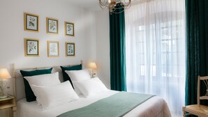 Ein ordentlich bezogenes Bett mit weißer und grüner Bettwäsche, eine Chandelüster, gerahmte botanische Drucke an der Wand und ein Fenster mit durchsichtigen Vorhängen.