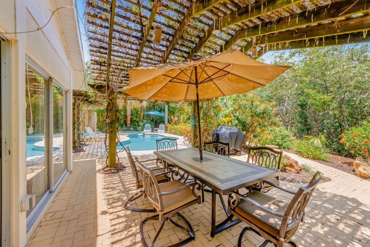 Een terras buiten met een tafel en stoelen, een pergola erboven en een zwembad op de achtergrond.