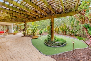 A covered outdoor area with a brick floor, surrounded by lush greenery and a small artificial turf area.
