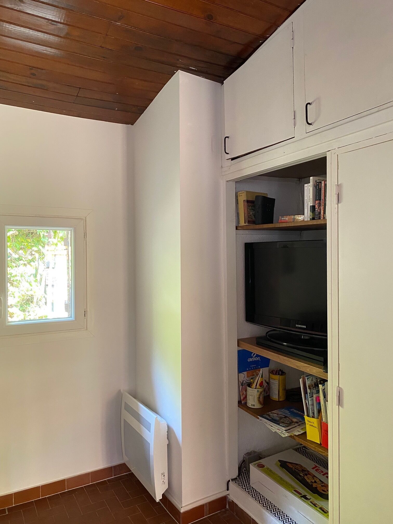 Een kamer met een witte radiator, een raam met uitzicht op groen, een wit kastje met een televisie, en een plank met boeken en schrijfwaren.