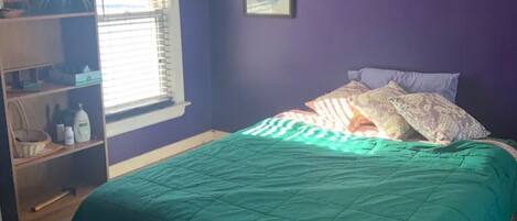 Un dormitorio con colcha color turquesa, una ventana con persianas, un estante con diversos objetos y un cuadro enmarcado en la pared.