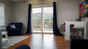 A spacious living room with a large sliding glass door leading to a balcony, a white desk, a blue armchair, and a small white table. - Vacation home you’ll enjoy (Lower Sackville)