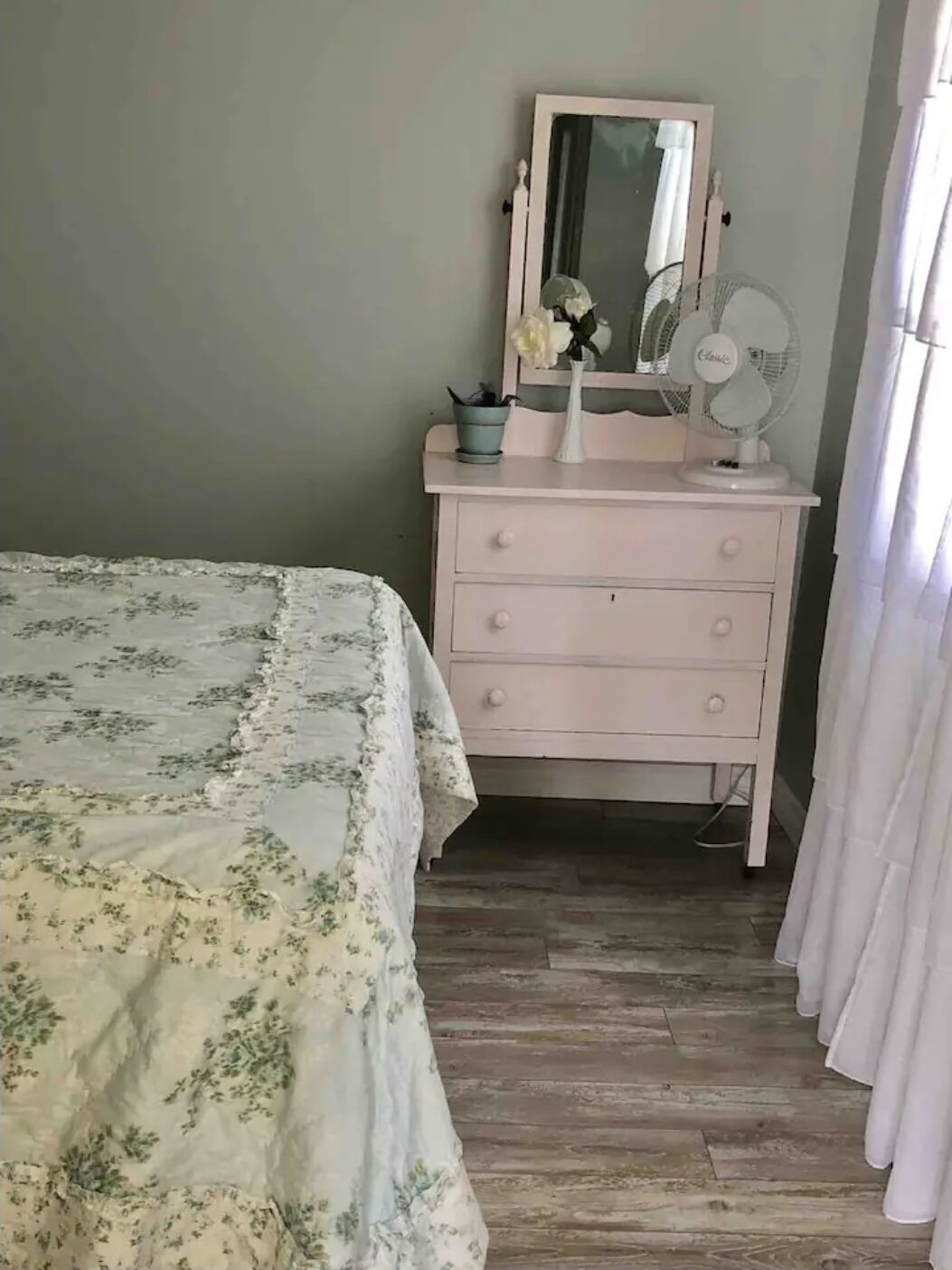 A bedroom with a bed, a dresser, a mirror, a fan, and a window with curtains.