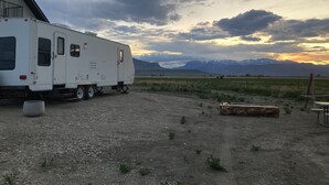 A white RV parked in a gravel area with a mountainous backdrop and a cloudy sky. - Camper/RV @ The Rendezvous - Wi-Fi, fire pit, A/C (Cody)