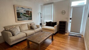 Un soggiorno con un divano beige, un tavolino in legno, un quadro appeso al muro, una ventola a soffitto e una porta con una finestra.