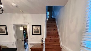 Una scala in legno conduce a un secondo piano, dove si trova una sala da pranzo con tavolo e sedie, e alle pareti sono appese delle cornici con immagini.