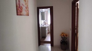 Un couloir avec un sol carrelé, un tableau accroché au mur, une plante en pot avec des fleurs et une porte donnant sur une salle de bain.