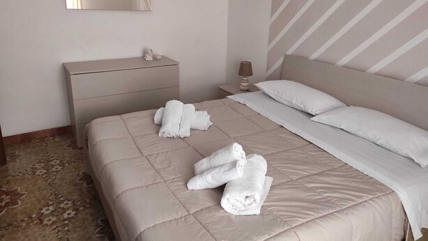 Un lit bien fait, avec du linge de lit blanc et des serviettes pliées, une table de chevet avec une lampe et une commode avec un miroir.