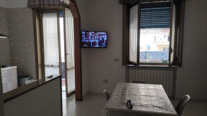 Une pièce comprenant un coin cuisine, une table à manger avec des chaises, un téléviseur fixé au mur et une fenêtre avec des stores.