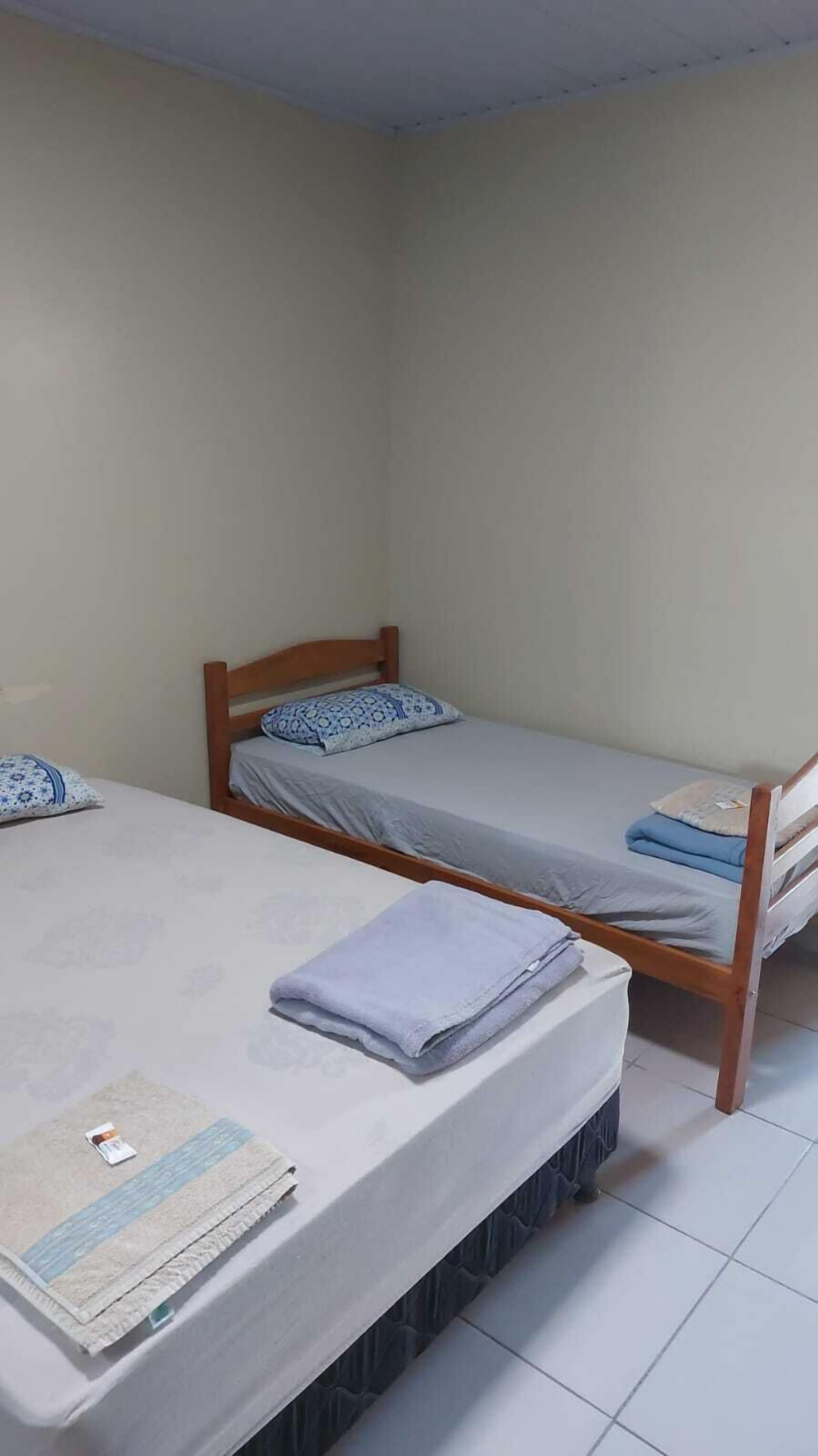 Una habitación con dos camas individuales, cada una con estructura de madera y colchón gris. Hay una mesita pequeña frente a las camas, con una toalla doblada y una revista.
