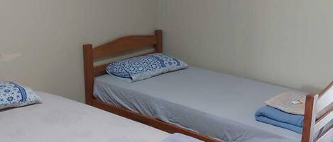 A room with two single beds, each with a wooden frame and a grey mattress. There is a small table in front of the beds with a folded towel and a magazine.