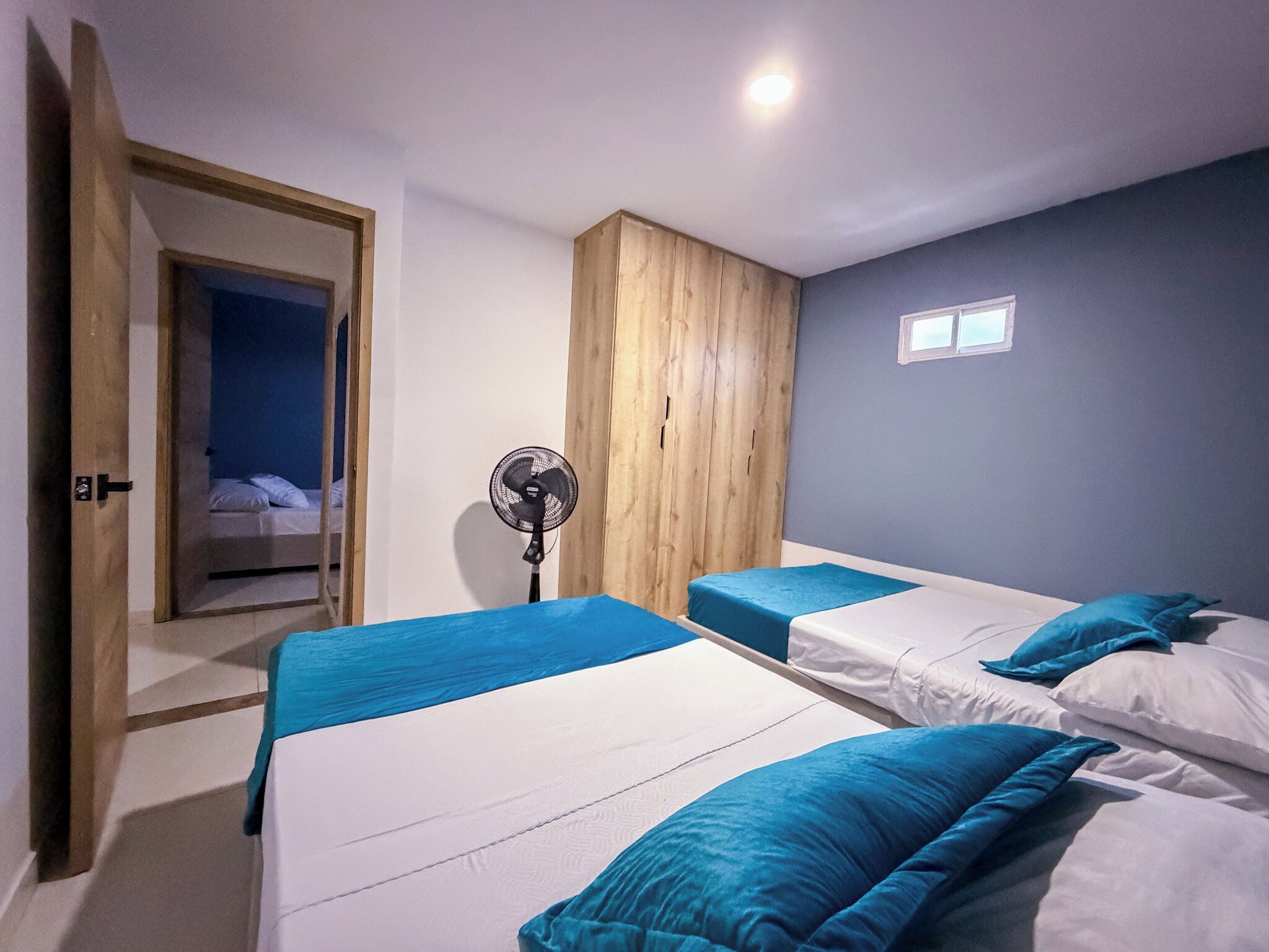Une chambre avec deux lits, un ventilateur sur pied, une armoire en bois et une porte donnant sur une autre pièce.