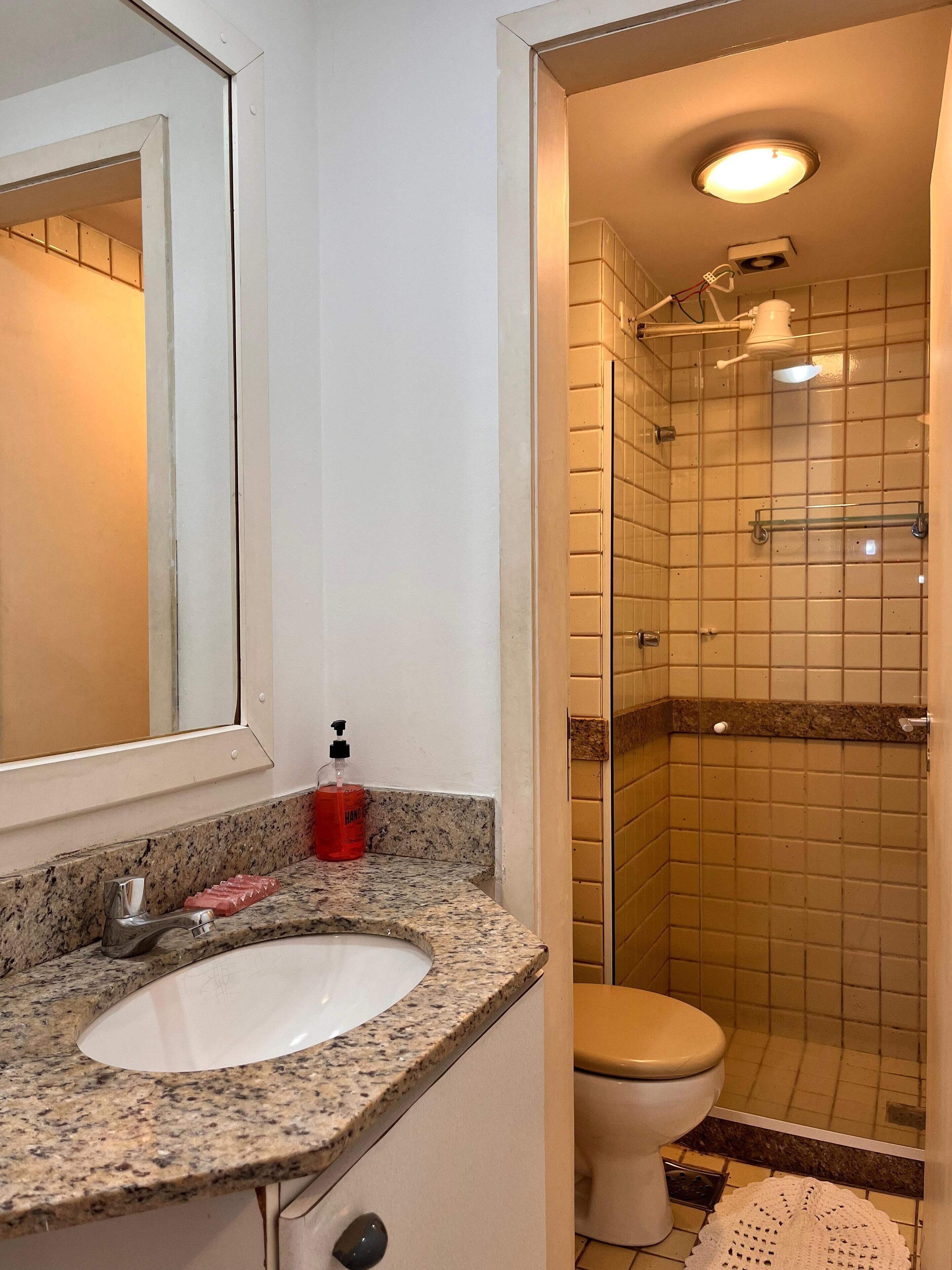 Un baño con lavamanos de encimera de granito, un espejo, un dispensador de jabón rojo, un inodoro y una zona de ducha con paredes de azulejos y una lámpara en el techo.