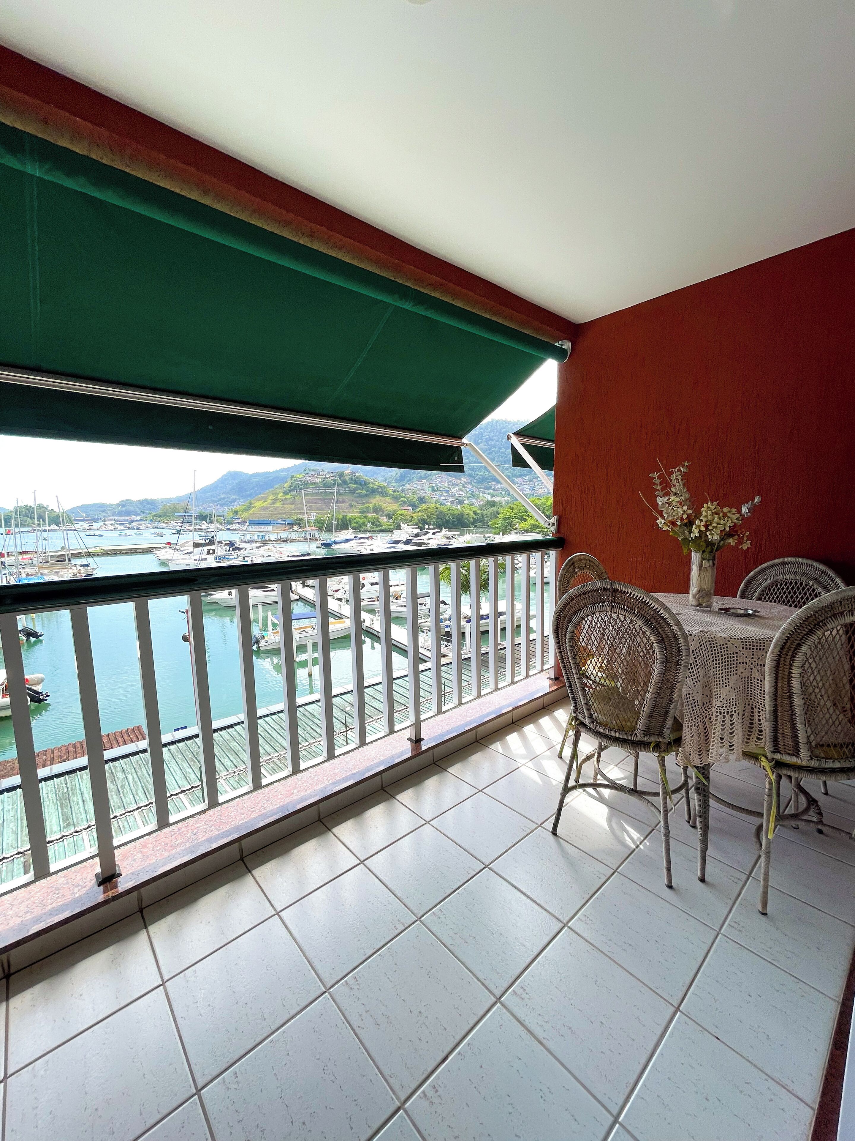 A balcony with a view of a marina and mountains. There is a table with chairs and a vase of flowers.