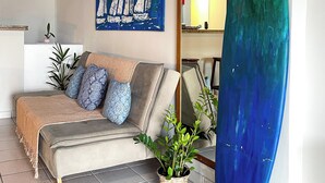 Una sala de estar con sofá, un cuadro de veleros, una tabla de surf y una planta en maceta.