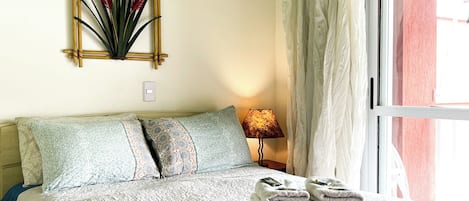Una cama bien hecha con una manta texturizada, una planta decorativa en la pared, una lámpara de noche y una ventana con cortinas.
