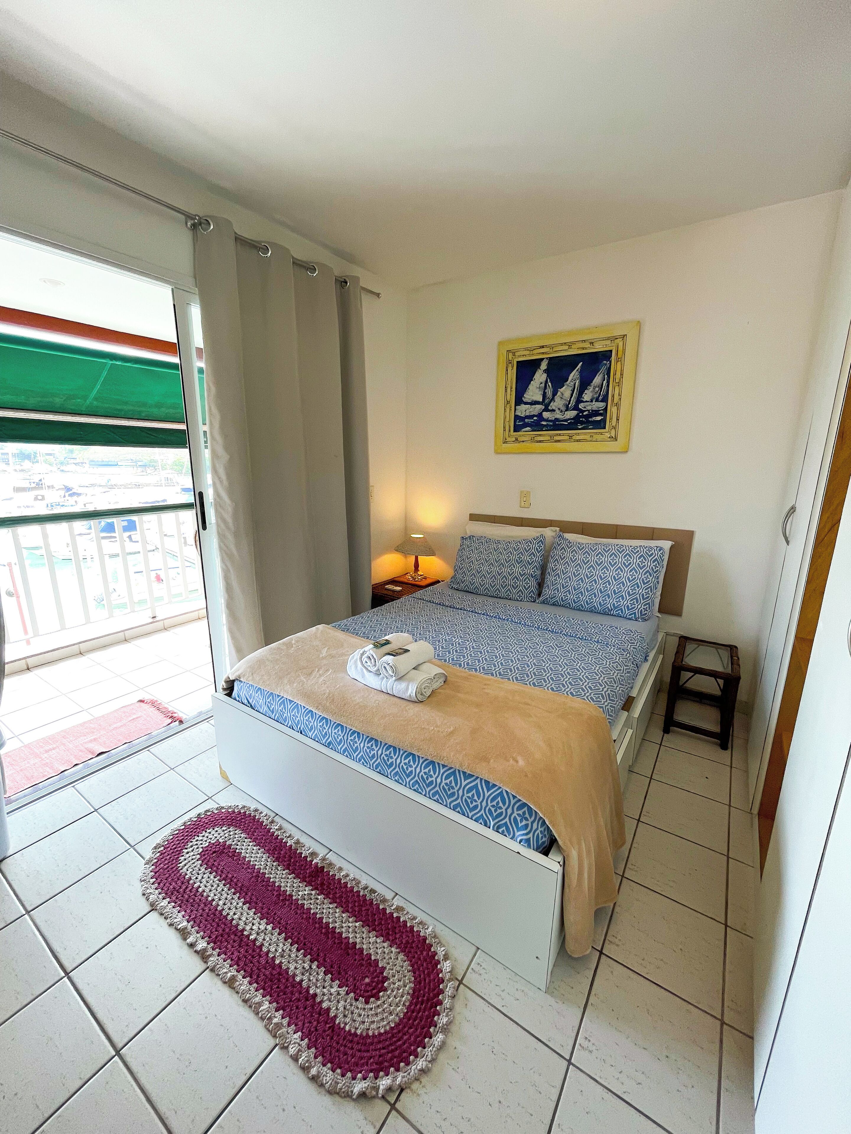 Une chambre à coucher avec un lit, un petit tabouret, une table de chevet, une lampe, un tableau et une vue sur l’extérieur par une fenêtre.