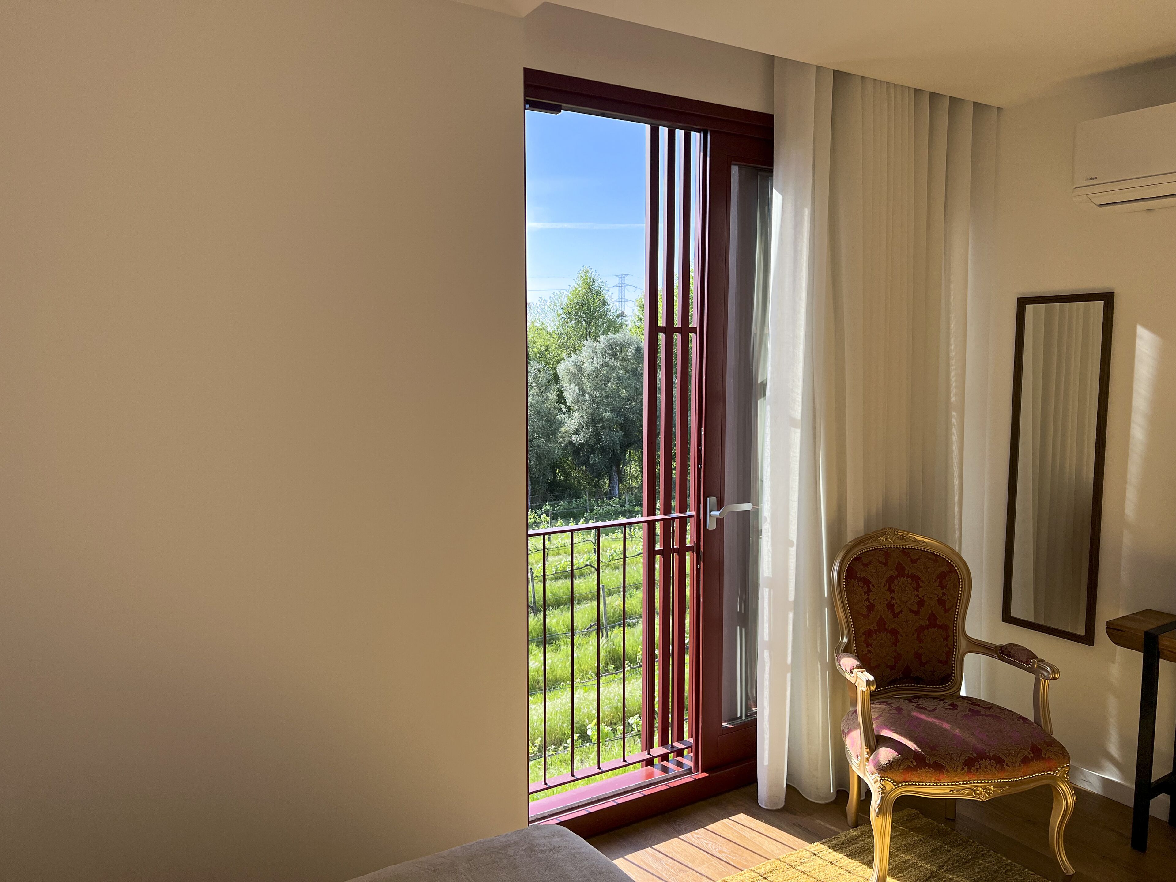 A room with a large sliding glass door leading to a balcony with a view of greenery and trees. There is a chair with ornate detailing and a small table beside it.