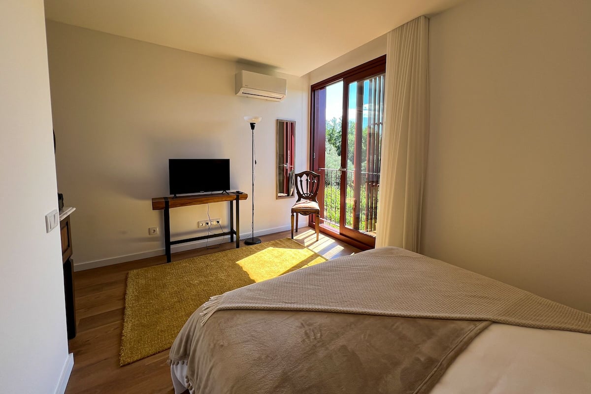 Een slaapkamer met een bed, een televisie op een houten standaard, een stoel bij het raam en een balkon met uitzicht.