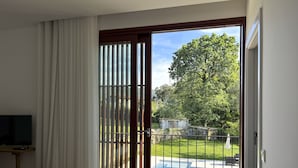 Une chambre avec un lit, une télévision, une table en bois et une vue sur une piscine et un jardin, accessible par une porte-fenêtre coulissante.