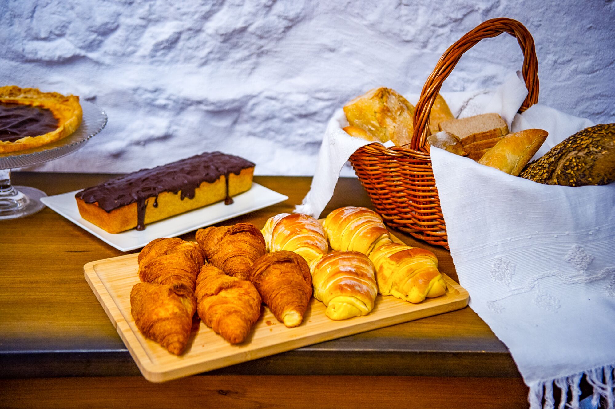 A wooden table with pastries, including croissants and a chocolate-topped cake, and a wicker basket with bread.