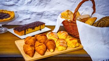 A wooden table with pastries, including croissants and a chocolate-topped cake, and a wicker basket with bread.