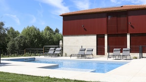 Une piscine avec des chaises longues, un bâtiment rouge aux grandes fenêtres et un ciel bleu azur.