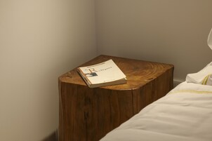 A neatly made bed with white bedding and a single book titled "R" on a wooden nightstand.