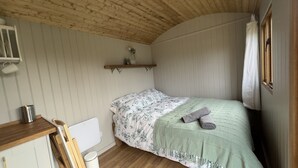 A cozy bedroom with a bed, a wooden desk, a white radiator, and a shelf with decorative items. - Luxury Shepherds Hut, W/large Garden, Firepit, & Wi-fi @ Cedar Gables Campsite (Flimwell, Wadhurst)
