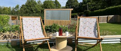 Two wooden chairs with patterned fabric, a small round table with a red mug and a white mug, and a metal shed in the background.