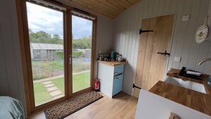 A compact kitchen with a sliding glass door leading to a garden, a small refrigerator, and a wooden countertop. - Luxury Shepherds Hut, W/large Garden, Firepit, & Wi-fi @ Cedar Gables Campsite (Flimwell, Wadhurst)